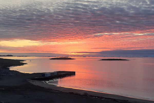 The still waters of a calm bay reflect the pink-red skies overhead.