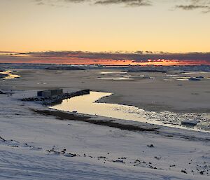 The same view as photograph 1 (Davis Beach) but this time covered in snow. The sea is mostly frozen and is also covered in snow. The sun has set in the distance and the sky has a red hue.