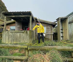 A person in a yellow jacket standing in front of a field hut, surrounded by lush grass