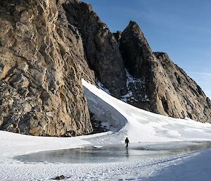 A person is waking across a frozen lake in front of a rocky mountain