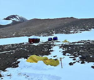 Three people are sleeping in yellow bags in the snow near a vehicle and hut