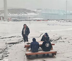 A woman and three men are gathered at a picnic table in the snow