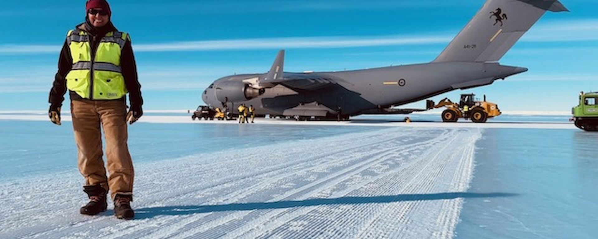 Person in winter clothing and hi-vis vest standing in front of a large Airforce cargo plane, on a runway made of ice.