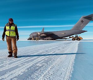 Person in winter clothing and hi-vis vest standing in front of a large Airforce cargo plane, on a runway made of ice.