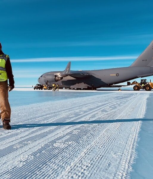 Person in winter clothing and hi-vis vest standing in front of a large Airforce cargo plane, on a runway made of ice.