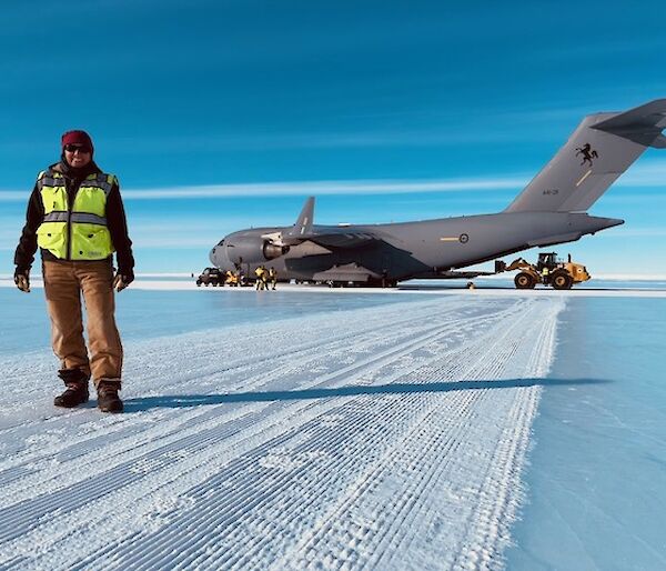 Person in winter clothing and hi-vis vest standing in front of a large Airforce cargo plane, on a runway made of ice.