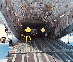 Looking into the rear of a large Airforce cargo plane that has a wide ramp lowered to unload cargo. A person with their back to the camera is standing on the ramp and guiding a vehicle that is being unloaded.