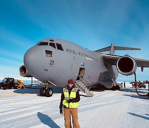 Person in winter clothing and hi-vis vest standing in front of the nose of a large Airforce cargo plane, on a runway made of ice. The aircraft is being refuelled, and there is a fuelling hose leading up into the wing.