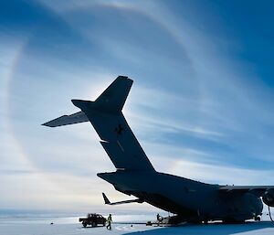 A large Airforce cargo plane is parked on a large flat area of ice. The view is from the rear right hand side of the aircraft, and a very large halo of sunshine is circling the tail of the plane, framing it. There are light wispy clouds in the blue sky, and the sun is being refracted through them in a circle shape.