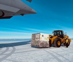 A large yellow front end loader is parked on an ice runway, with a big white box on the front of it, ready to load into an Airforce cargo plane. The very end of the aircraft only, is visible at the top left of the picture.