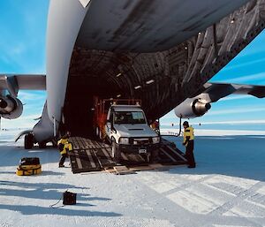 Looking into the rear of a large Airforce cargo plane that has a wide ramp lowered to unload cargo. There is a person either side of the ramp wearing yellow winter clothing, guiding down a vehicle that is being unloaded.