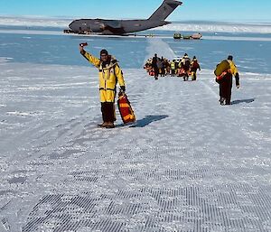 In the distant background, a large grey Airforce cargo plane sits on a runway made of ice. A bunched group of about 30 people, all dressed in yellow winter clothing walks towards the plane with their backs to the camera. In the foreground, one Expeditioner has turned to face the camera and has one arm raised high in the air, waving farewell to the photographer.