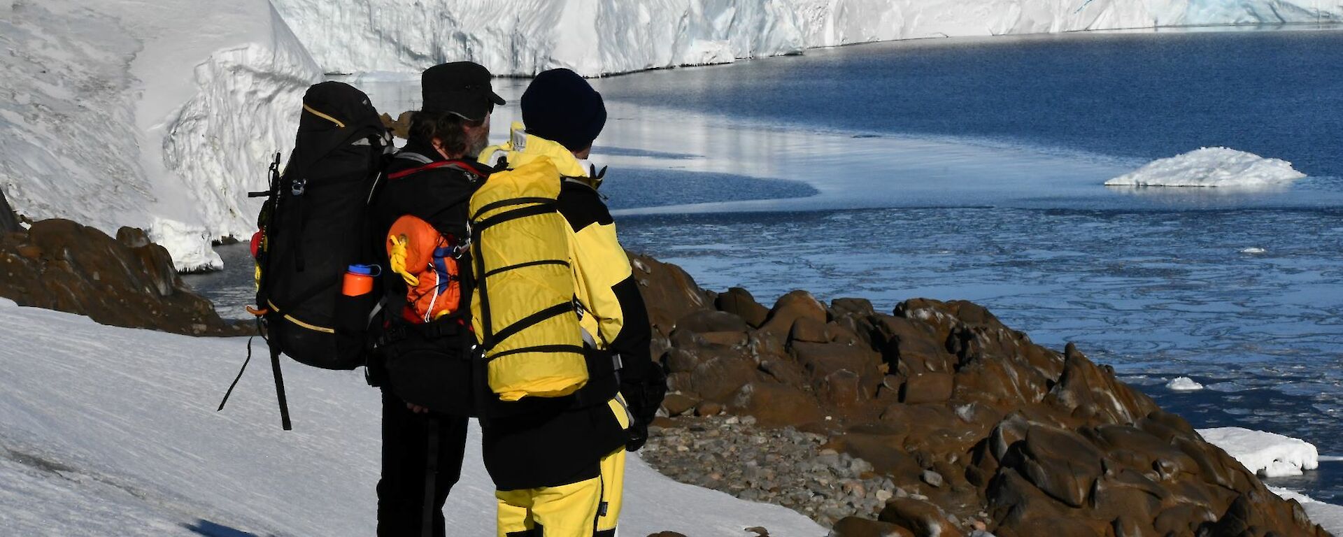 Two people stand on a snow covered hill, with the face of a large glacier stretching away in the background. The person on the right is wearing yellow winter pants and jacket, the person on the left has black clothing, and a large backpack. There are several large pieces of ice floating in the water in front of the crumbling glacier face.