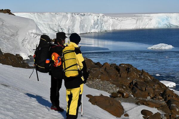 Two people stand on a snow covered hill, with the face of a large glacier stretching away in the background. The person on the right is wearing yellow winter pants and jacket, the person on the left has black clothing, and a large backpack. There are several large pieces of ice floating in the water in front of the crumbling glacier face.