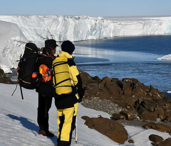 Two people stand on a snow covered hill, with the face of a large glacier stretching away in the background. The person on the right is wearing yellow winter pants and jacket, the person on the left has black clothing, and a large backpack. There are several large pieces of ice floating in the water in front of the crumbling glacier face.