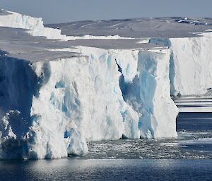 The photo shows a wall of white and light blue ice, stretching from left to right across the frame, with calm, flat dark blue water at its' base. The ice wall is cracked and fractured from top to bottom. The sun is reflecting the ice wall in the calm water.