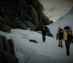 Three people, viewed from behind, are hiking along a snow covered path in mostly yellow winter survival clothing, with large backpacks. To the left of the people is a wall of large brown boulders, and to the right is a wall of ice and snow.