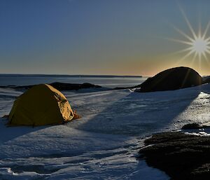 A tent set up on the snow with the sun low on the horizon