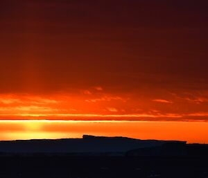 Clouds stretch across the frame from left to right, illuminated bright orange b y the setting sun, which is hidden behind the lower part of the cloud. In the lower third of the photo is the outline of a large iceberg, silhouetted black.