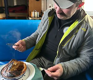 A person sits at a small table with knife and fork in hand, raised above a plate with a baked pie on it. The person is wearing a grey jacket and a grey hat, and is looking at the pie questioningly.