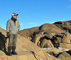 A person in a grey jacket and brown pants is standing on a large brown rocky outcrop. The person has sunglasses on and is looking out to the right of the photo thoughtfully, holding a camera in their right hand. The sky in the background is clear and sunny.