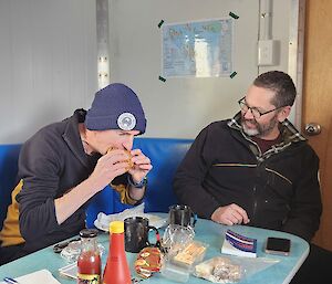 A person is sitting at a small table with a hamburger raised to their mouth, about to take a bite. The person is wearing a black jumper and a dark blue beanie. Another person sits to the right of them, laughing at the first person. This person is also wearing a black jumper, with no head wear, and has reading glasses on. The table is covered in cups, sauce bottles, and books.
