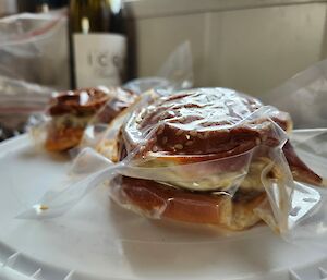 A hamburger in plastic wrap that has been shrunk down with a vacuum sealing machine sits on top of a white plastic container lid.