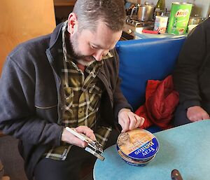 A person in a black jumper, and green flannelette shirt is sitting at a small table with a tinned meat pie in front of them. The tin is blue and says Fray Bentos. The person is laughing and holding a can opener in their right hand. In the background is a small kitchenette with a tin of Milo on the bench.