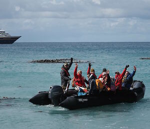 People in an inflatable boat waving to the camera, with a cruise ship in the background