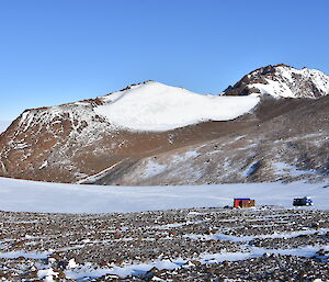 A snow covered mountain range rises behind a blue vehicle and red hut