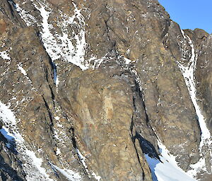 A hut,vehicle and two people are dwarfed by a rocky mountain