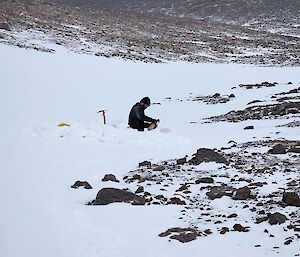 A man is standing in a hole in the snow with an ice axe next to him