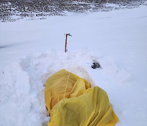 A yellow bivvy bag lays in a hole in the snow with an ice axe behind