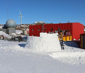 A man is working on building the wall of an igloo. A red building, wind turbine and satellite dome are in the background