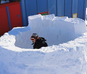 A man wearing googles and a beanie is crouched in the middle of the first stage of an igloo