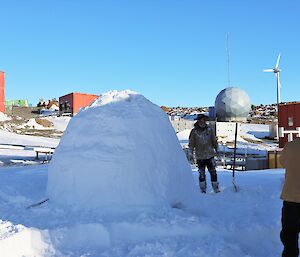 A man stands next to an igloo with buildings, a satellite dome and wind turbine in the background