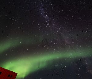 An aurora is visible in the sky with the milky way and a red building in the bottom corner