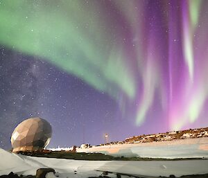 An aurora of pink, purple and green and the milky way are in the sky above a satellite dome.