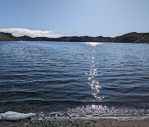 A view from the shoreline of a lake looking out over glistening water surrounded by rocky hills.