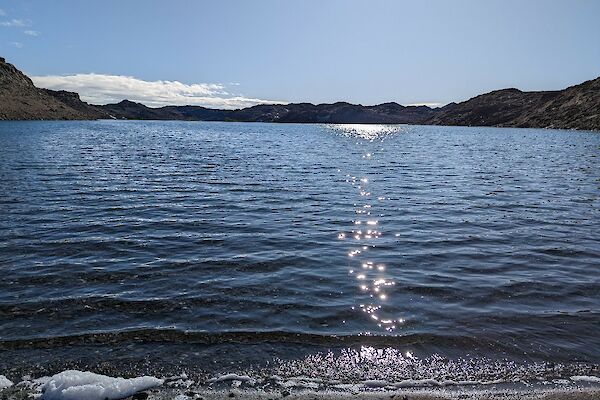 A view from the shoreline of a lake looking out over glistening water surrounded by rocky hills.
