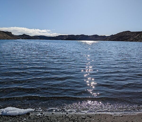 A view from the shoreline of a lake looking out over glistening water surrounded by rocky hills.