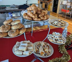 Plates of bagels, croissants, fruit mince tarts and assorted pastries on a table covered in a red tablecloth.
