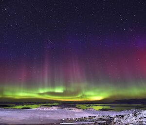 The foreground of the photo is a thin layer of snow. It is night time but the sky is covered in light. Firstly a base of wavy light green, with red highlights. Out of this wavy base there are vertical beams stretching up into the star filled sky. The beams are green, then pink, then purple, finally transitioning into the black night sky. The water of the bay beneath is lit up green with the reflection of the light in the sky, and is covered in small icebergs.