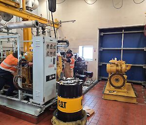 Two mechanics in Hi-vis workwear, are working behind a large grey electrical panel to remove an alternator from a yellow generator. There is a 44 Gallon drum of oil in the foreground with the work CAT on it.