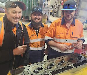 Three people in hi-vis work-wear are standing behind a long engine block, in the process of rebuilding it. The head of the engine has been removed and you can see the holes where the valves operate.