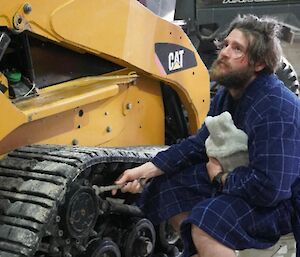 A scruffy looking person in a blue dressing gown and clutching a hot water bottle under one arm is crouched down in front of a skid steer loader (bobcat). They are holding a spanner and looking sadly into the distance.