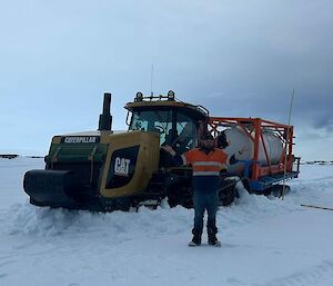 A person in Hi-vis workwear is standing in front of a large tractor that is towing a trailer with a fuel tank on it. The person has their arms raised up slightly. The tractor is stuck in a heavy layer of snow lying on the ground. The sky is completely overcast.