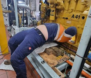 A person in HI-vis workwear is bent backwards, lying across a large piece of equipment, with knees bent. Their head is obscured underneath a large engine, changing out some bearings. The persons stomach is exposed under the bottom of their shirt.