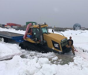 A large yellow tractor with an empty flatbed trailer is stuck in a layer of thick snow and ice, up to the top of it's tracks. There is a person in brown overalls and a black long sleeve shirt leaning against the front of the tractor, holding one leg up on the bumper of the tractor. The sky in the background is completely overcast.