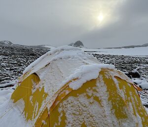 A yellow tent covered in snow as the sun tries to peak through the cloud cover